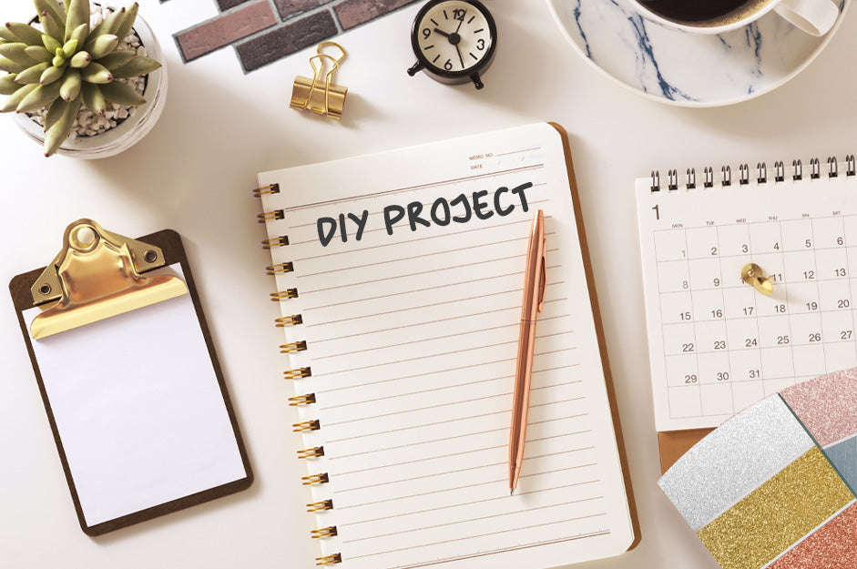 Flatlay with notebook in middle with text saying DIY PROJECT, circled of green flower pot, cup of coffee, vinyl samples and stationary
