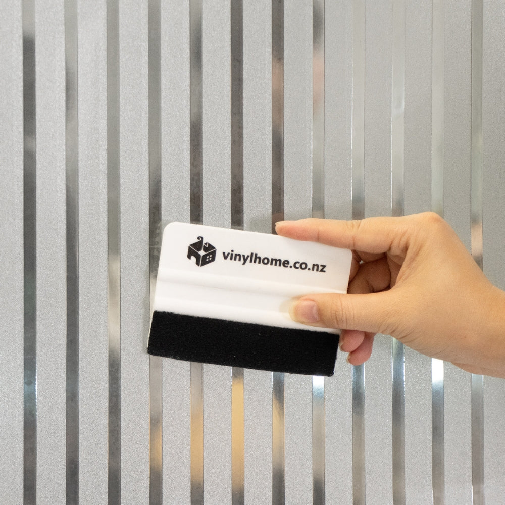 Hand holding a squeegee with 'vinylhome.co.nz' branding against a striped frosted window film
