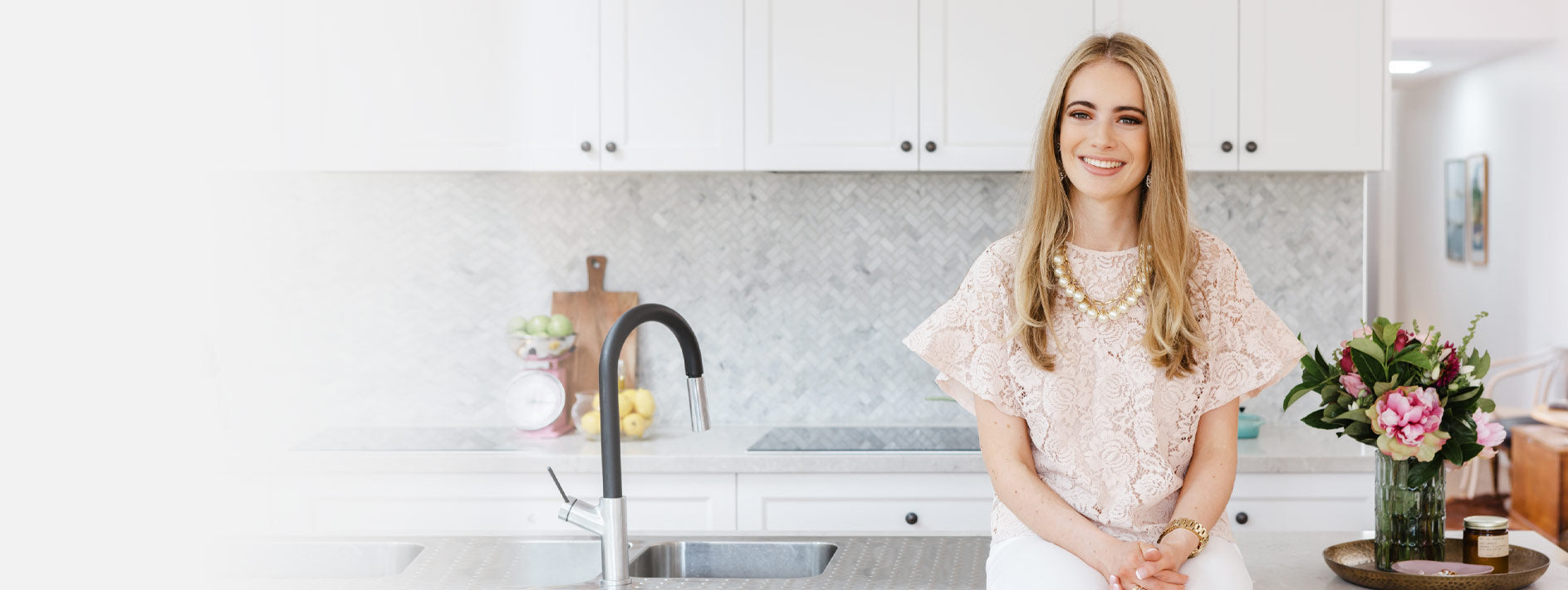 Blonde girl in pink top sitting on a kitchen island in front of a white kitchen with marble wall tiles