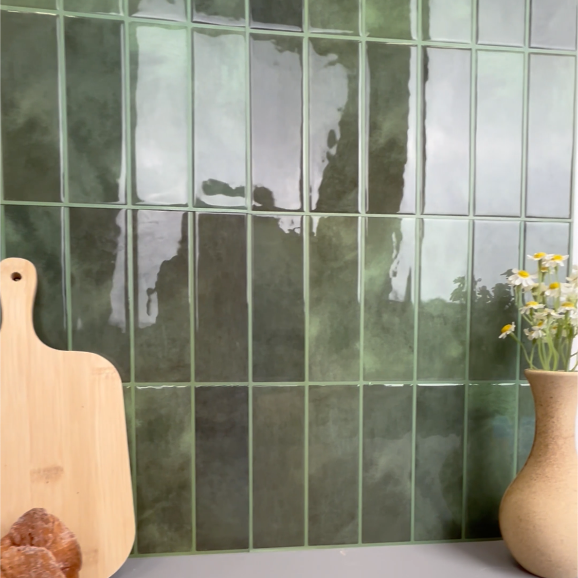 Green tiled wall with a wooden cutting board, bread, and vase on a gray surface.