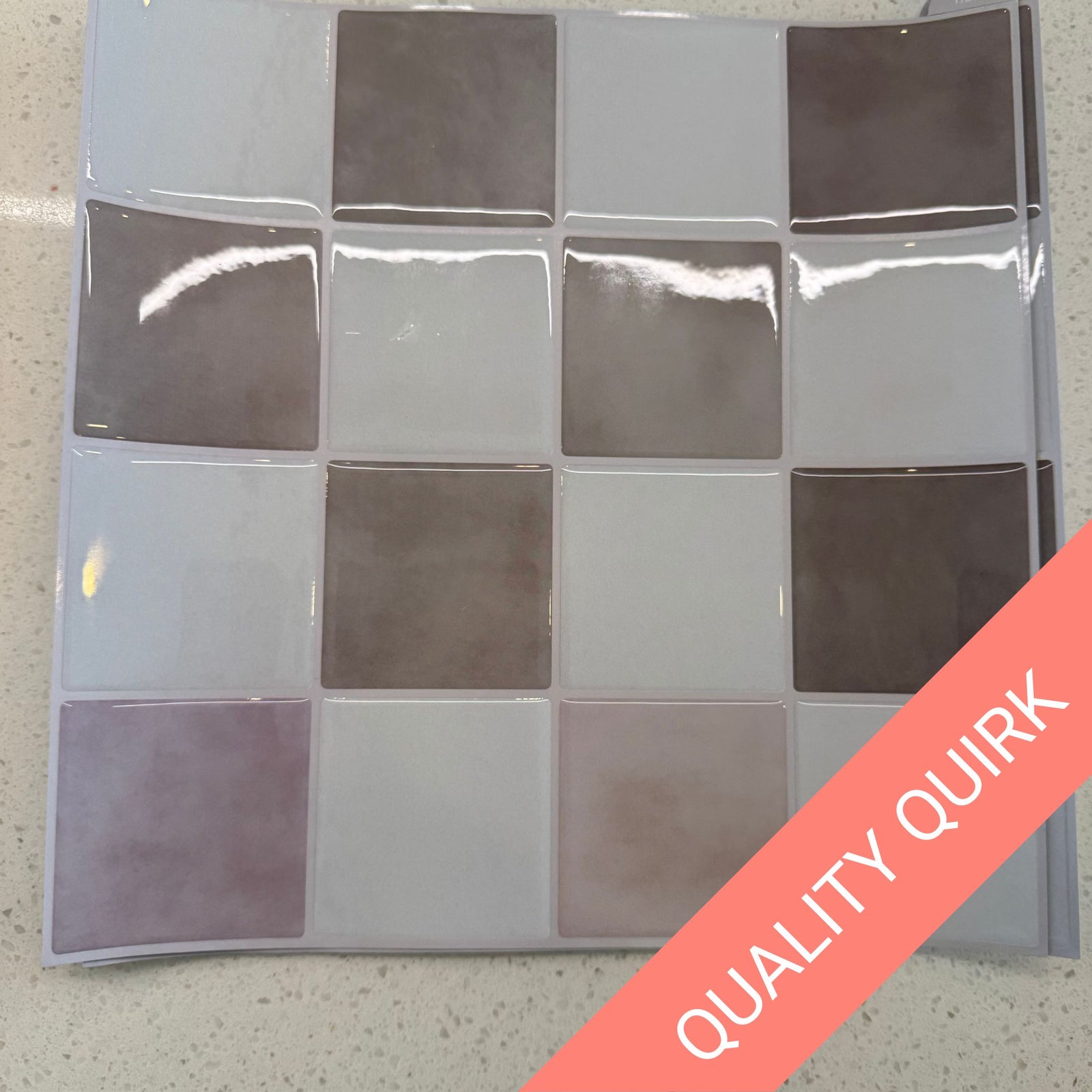 A glossy square tile sheet with a grid of 12 tiles in alternating light gray, white, and taupe-brown shades, shown on a speckled countertop with a coral diagonal banner reading 'QUALITY QUIRK'.