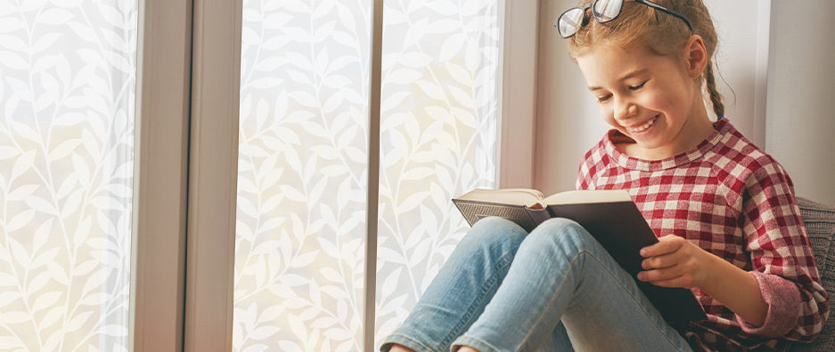Young girl with blonde braids, jeans and red white plaided shirt sitting in a window with leafy pattern window film reading a book