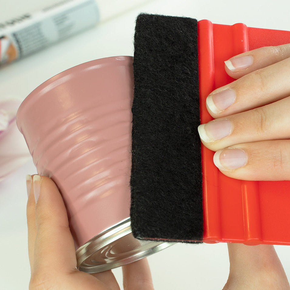 A close-up of hands holding a pink metal can with a black and red squeegee tool pressed against it, with a tube in the blurred background.