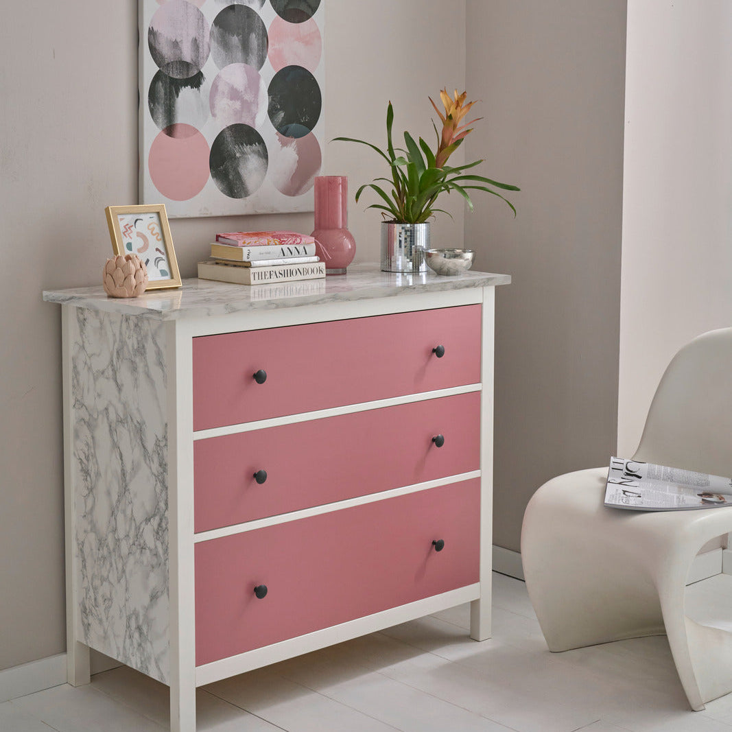 A three-drawer dresser with pink fronts and a white-and-grey marble-look side panel, styled in a minimalist room with a vase of flowers, books, and a framed print.