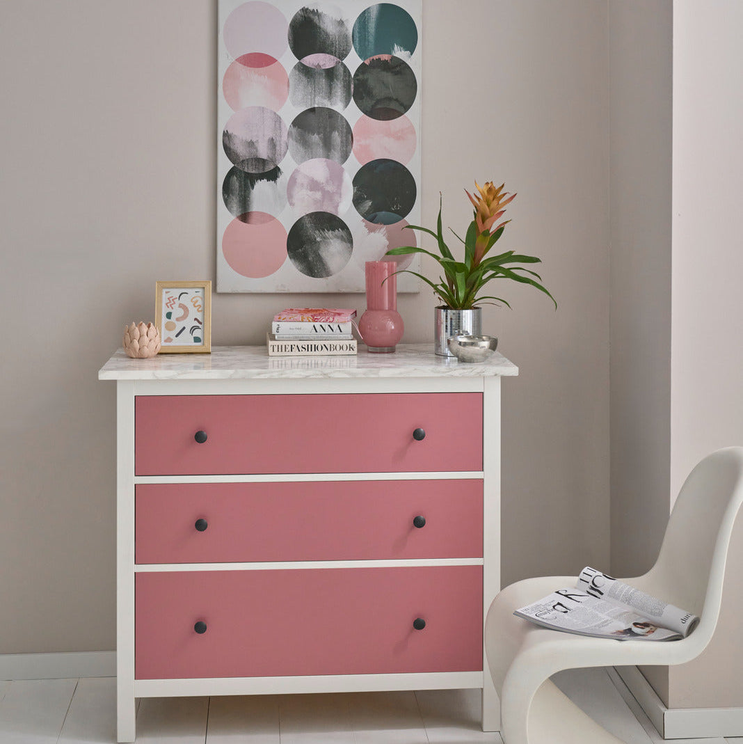 A white wooden dresser with three pink drawer fronts and small black knobs, styled in a room setting with a plant, books, and a framed print, next to a modern white chair.