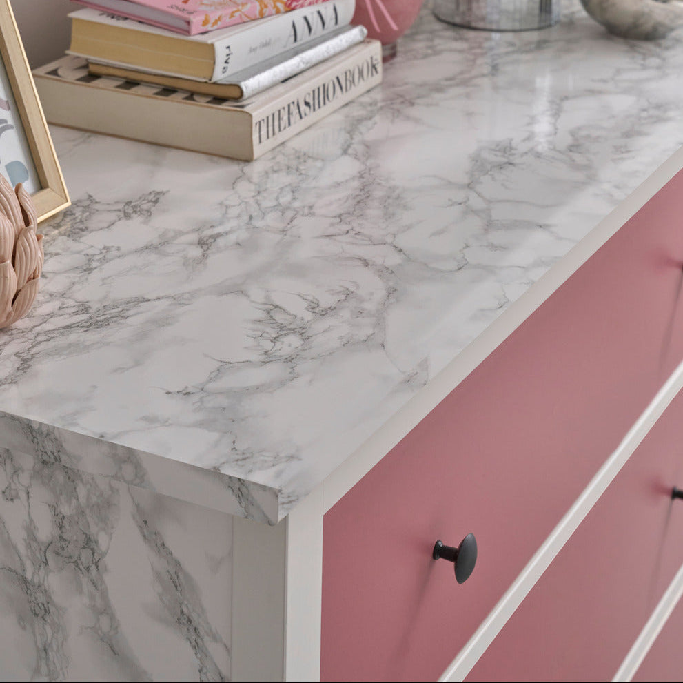 A pink and white marble-look dresser with pink drawer fronts, styled with books, a pink vase, a potted plant, and a small bowl in a bright interior setting.