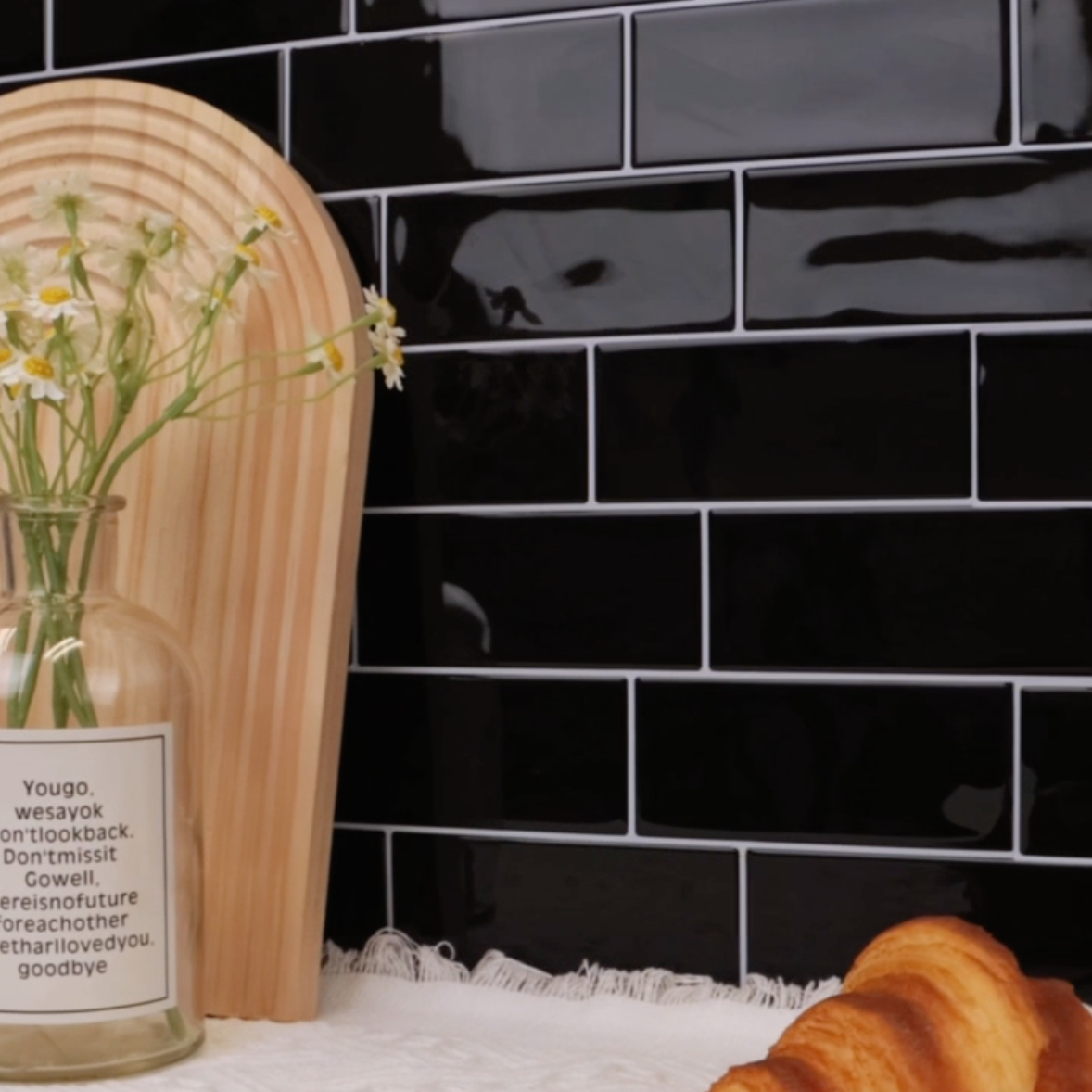 Black tiled wall with a croissant and a vase with flowers on a white surface.