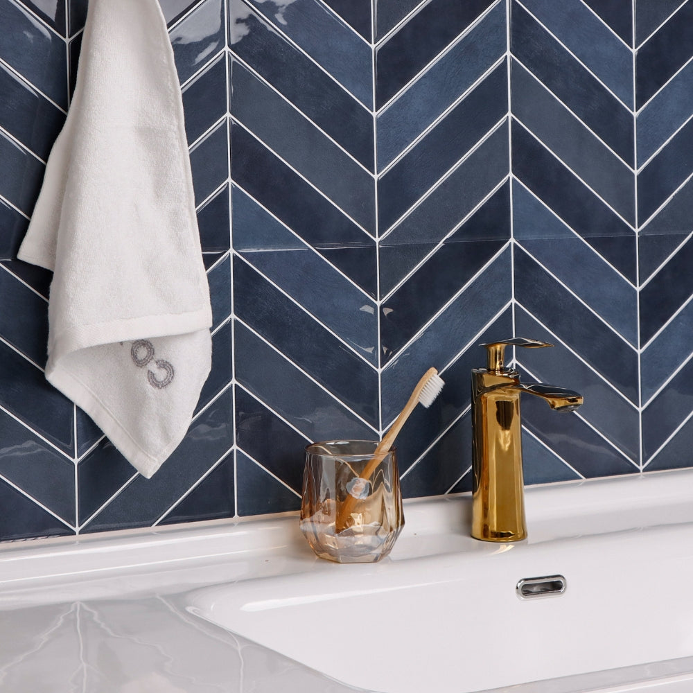 Bathroom sink area with gold faucet, glass, toothbrush, and towel against a blue chevron tiled wall.