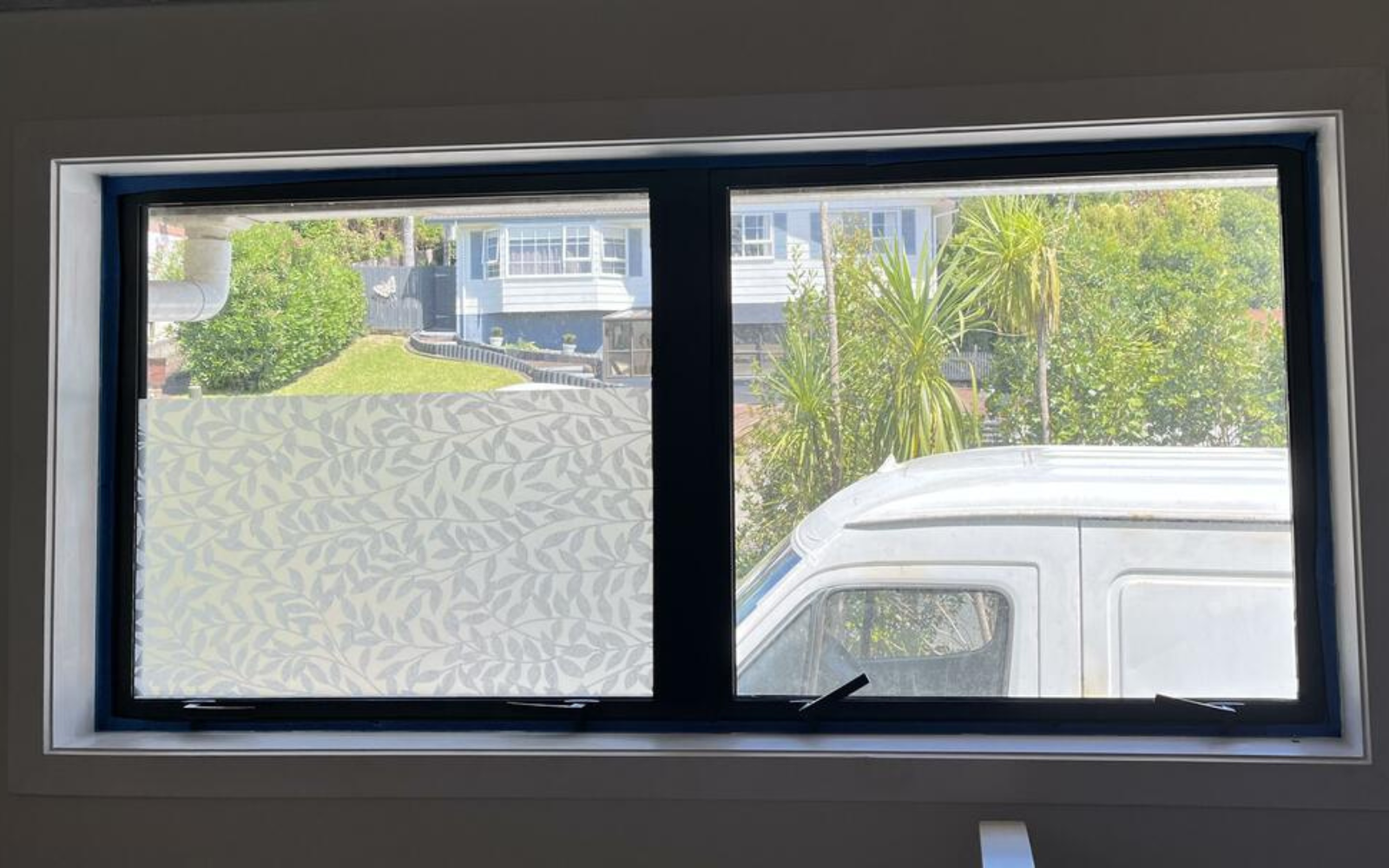Window with decorative film on one pane, showing a view of a garden and white van outside.