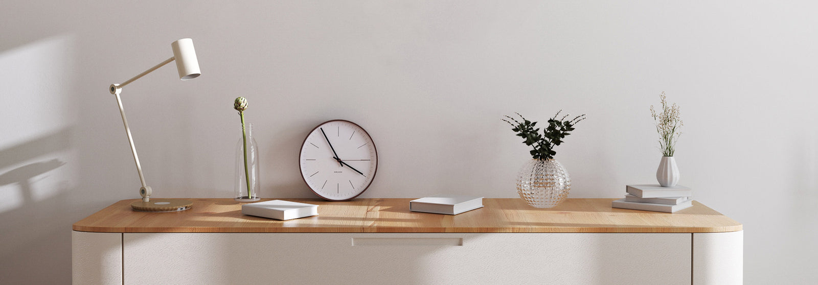 Oak top wooden dest with white metal lamp, clock and flowers on top, cream wall at the back