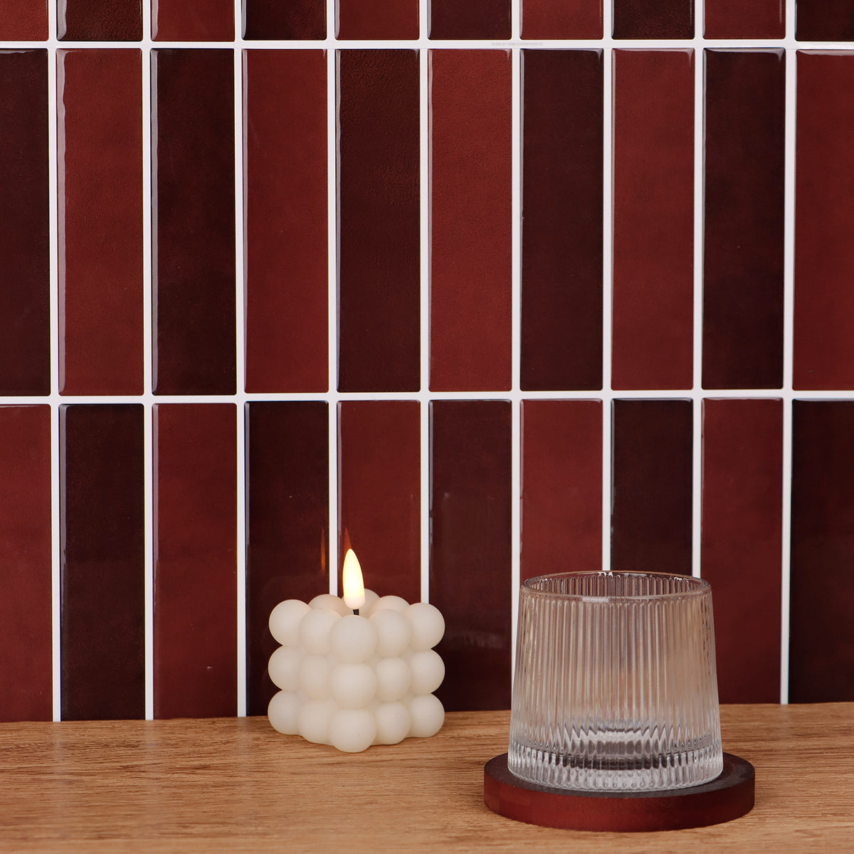 Two candles, one white textured and one clear glass, on a wooden surface with a tiled wall background.