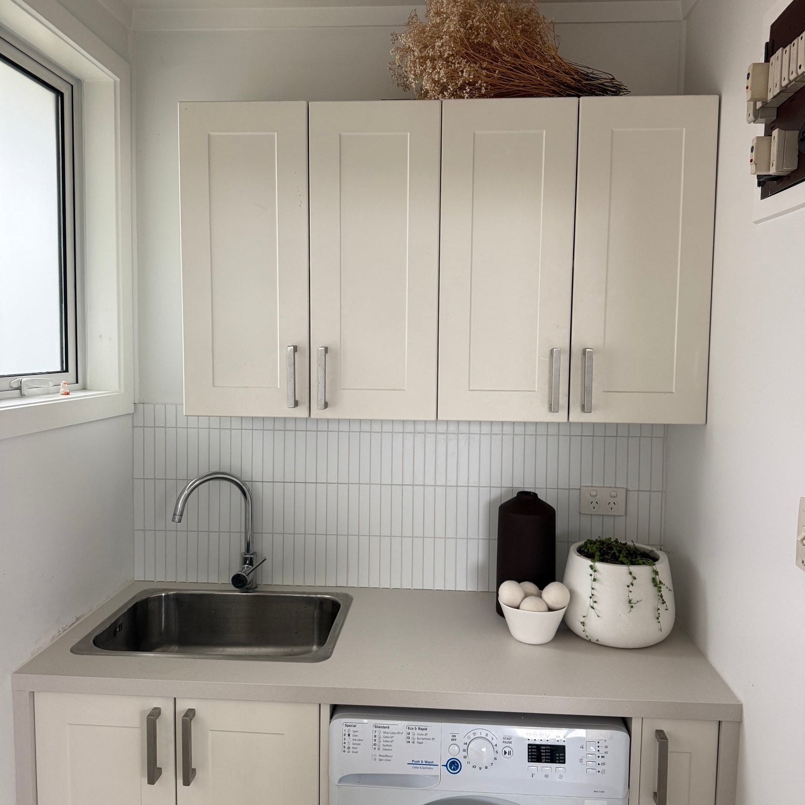 A compact utility room featuring cream-colored shaker-style cabinets, a stainless steel sink, a built-in front-loading washing machine, and a white matte kit kat tile backsplash.