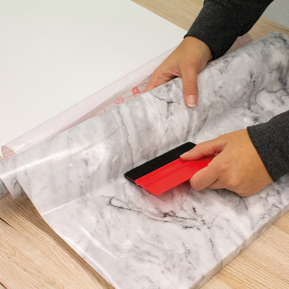 Person applying marble-patterned contact paper with a red tool on a white surface
