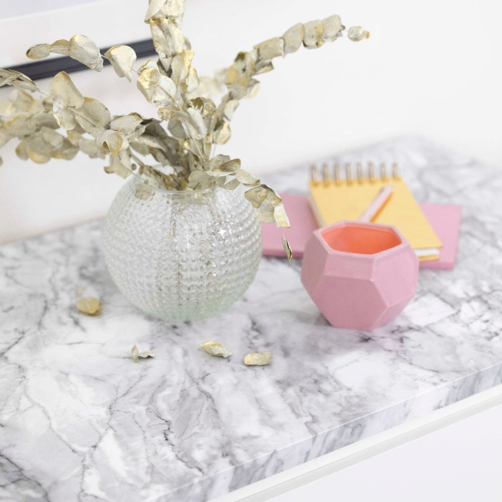 Decorative vase with gold leaves on a marble surface with a pink geometric cup and notebook.