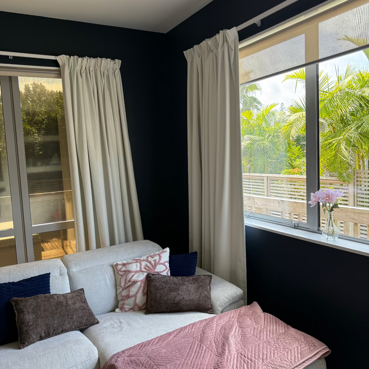 Living room with a white sofa, decorative pillows, and a pink blanket, featuring large windows with white curtains.