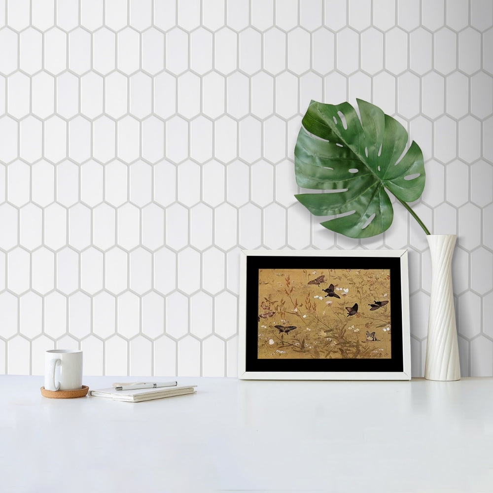 Framed artwork on a table with a plant and decorative items against a hexagonal tile wall.