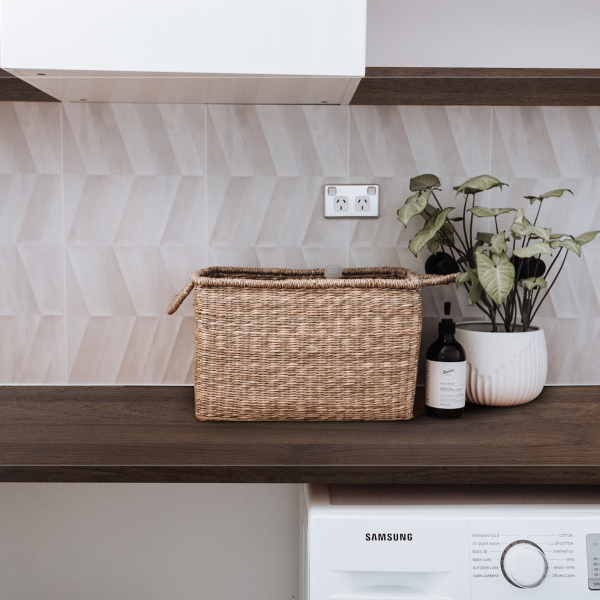 Laundry counter with a woven basket, plant, and bottle against a tiled wall.