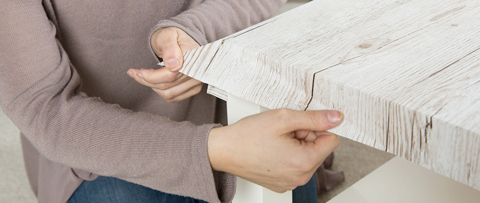 Close up of woman folding down wood grain self-adhesive vinyl over edge on coffee table