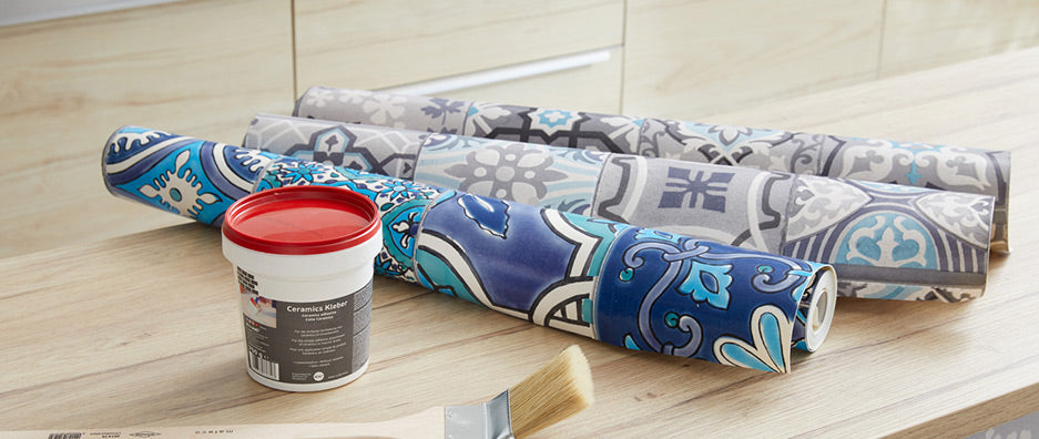 Moroccan design wallpaper rolls in blue, grey and mint on bench top together with white glue box and red lid
