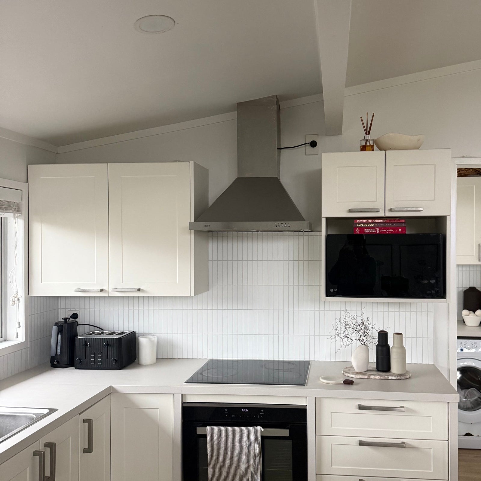 A modern kitchen interior with white cabinets, a black built-in oven and microwave, a stainless steel range hood, light wood flooring, and a small dog visible on the floor in the lower right corner.