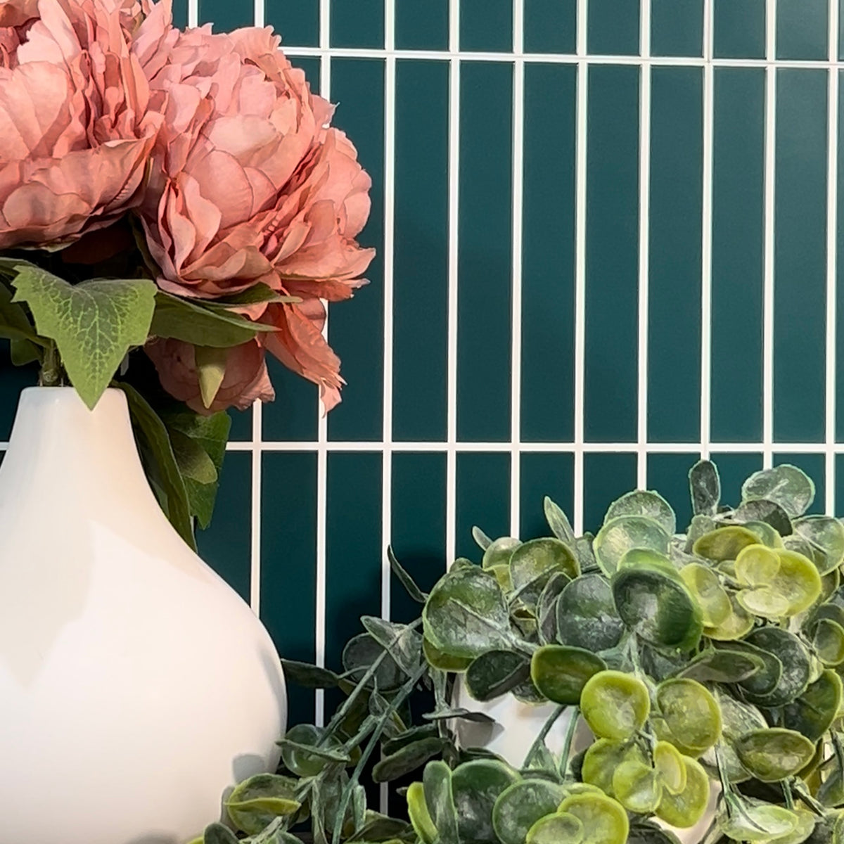 White vase with pink flowers and green foliage against a dark green tiled wall.