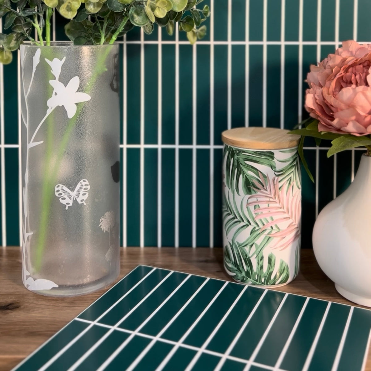 Decorative glass vase with floral etching, ceramic jar with leaf pattern, and white vase on a tiled surface.