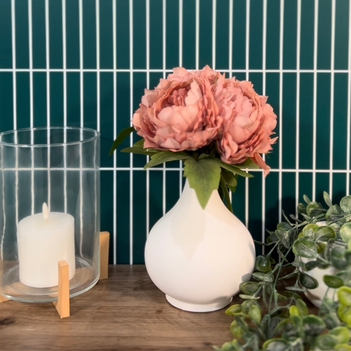 White vase with pink flowers on a wooden surface against a green tiled wall.