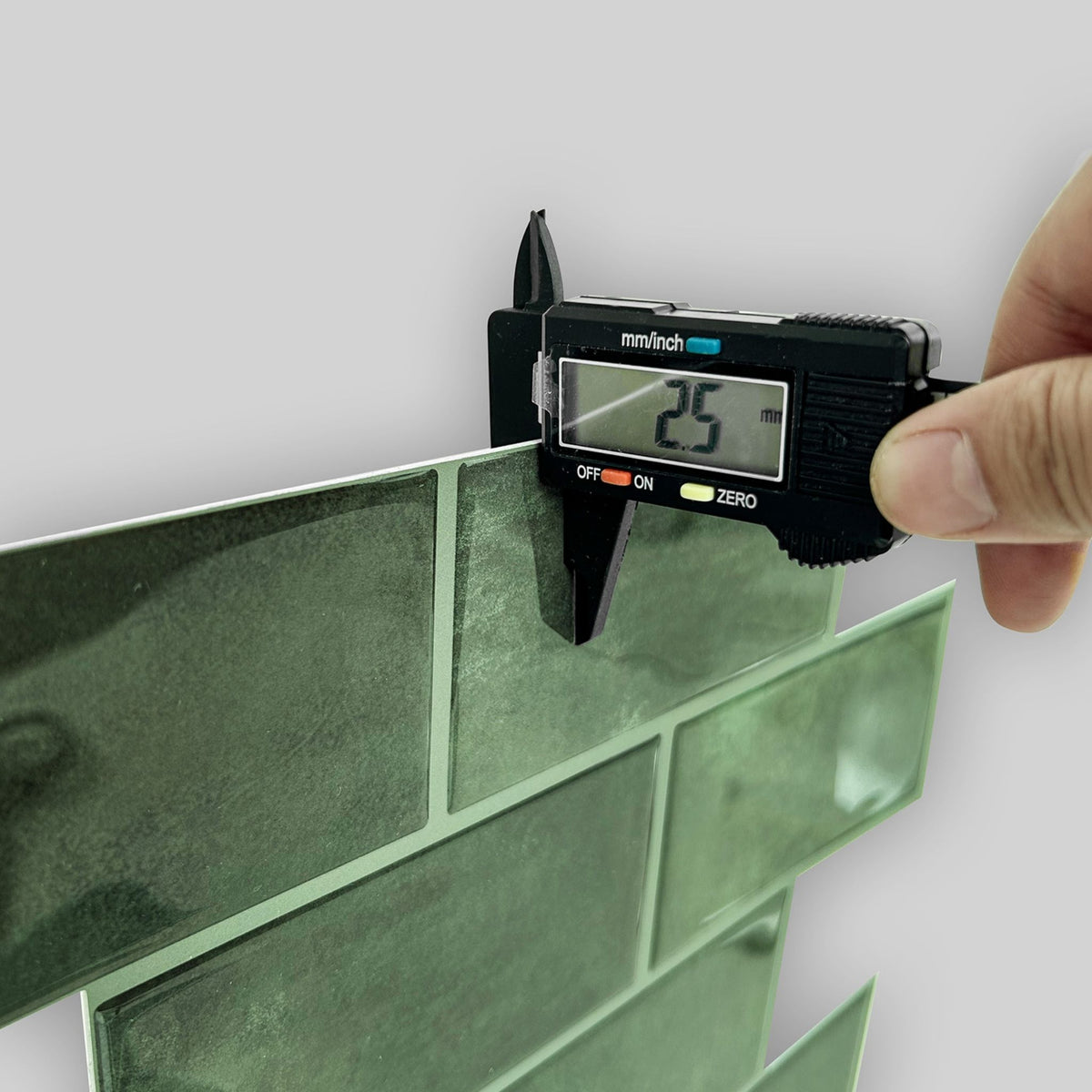 A hand holding a black digital caliper measuring the thickness of glossy green rectangular wall tiles against a light gray background.