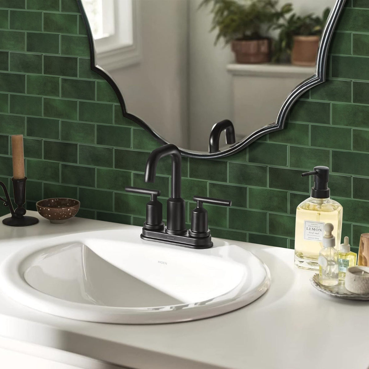 A bathroom vanity setup featuring a matte black two-handle faucet mounted on a white sink, set against a green subway tile wall with a decorative scalloped mirror and toiletries on the counter.
