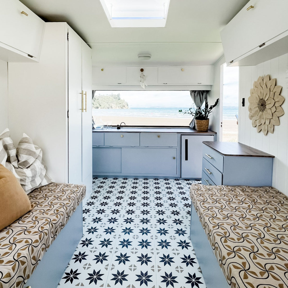 A compact beachside caravan interior with white walls and cabinets, pale blue kitchen cabinetry and side tables, a brown-and-cream star-pattern tile floor, patterned upholstered daybeds on both sides, and an ocean view through the back window.
