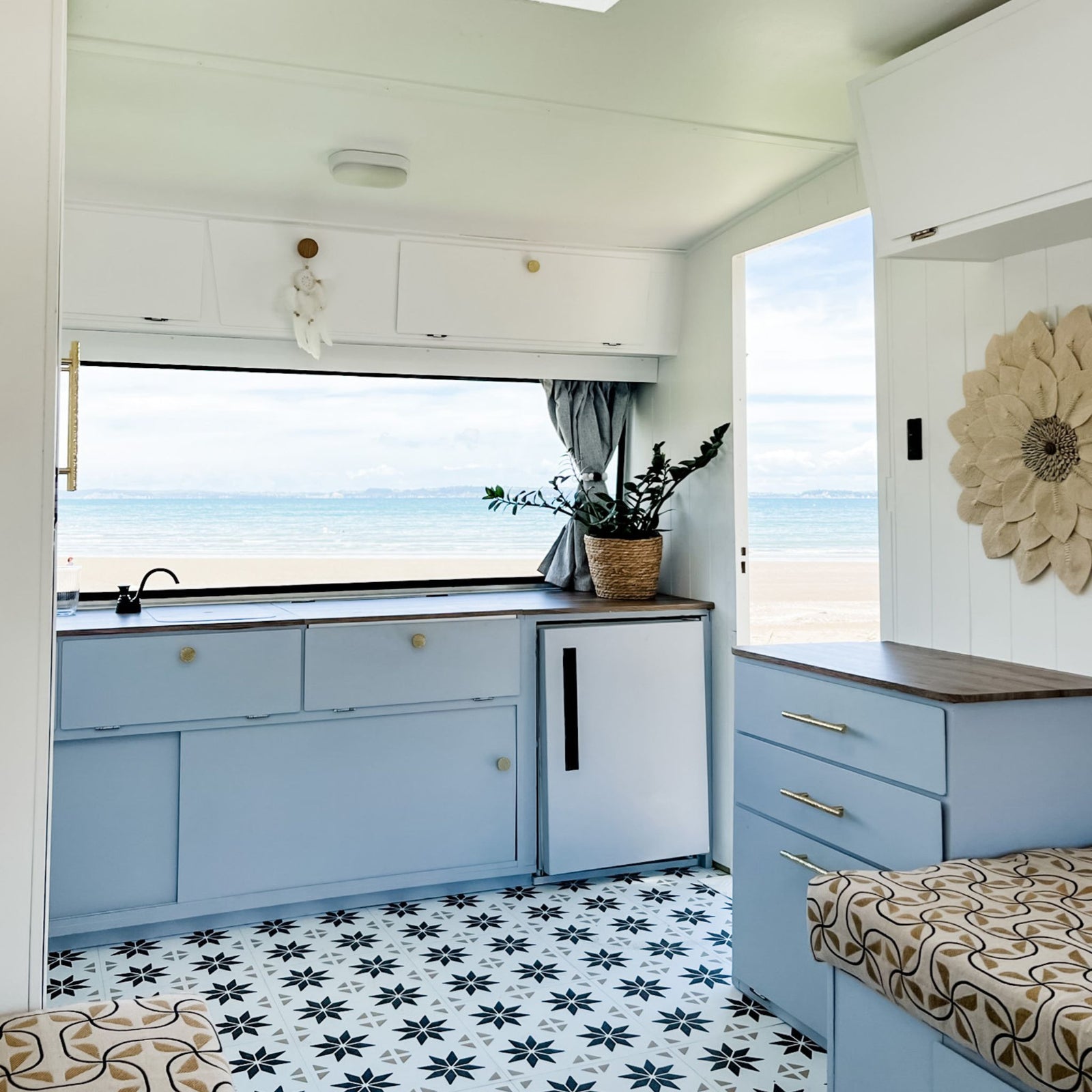 A compact coastal kitchenette inside a caravan, featuring light blue cabinets, a black countertop, a small white refrigerator, a patterned brown-and-beige floor tile, and a beach view through large windows.