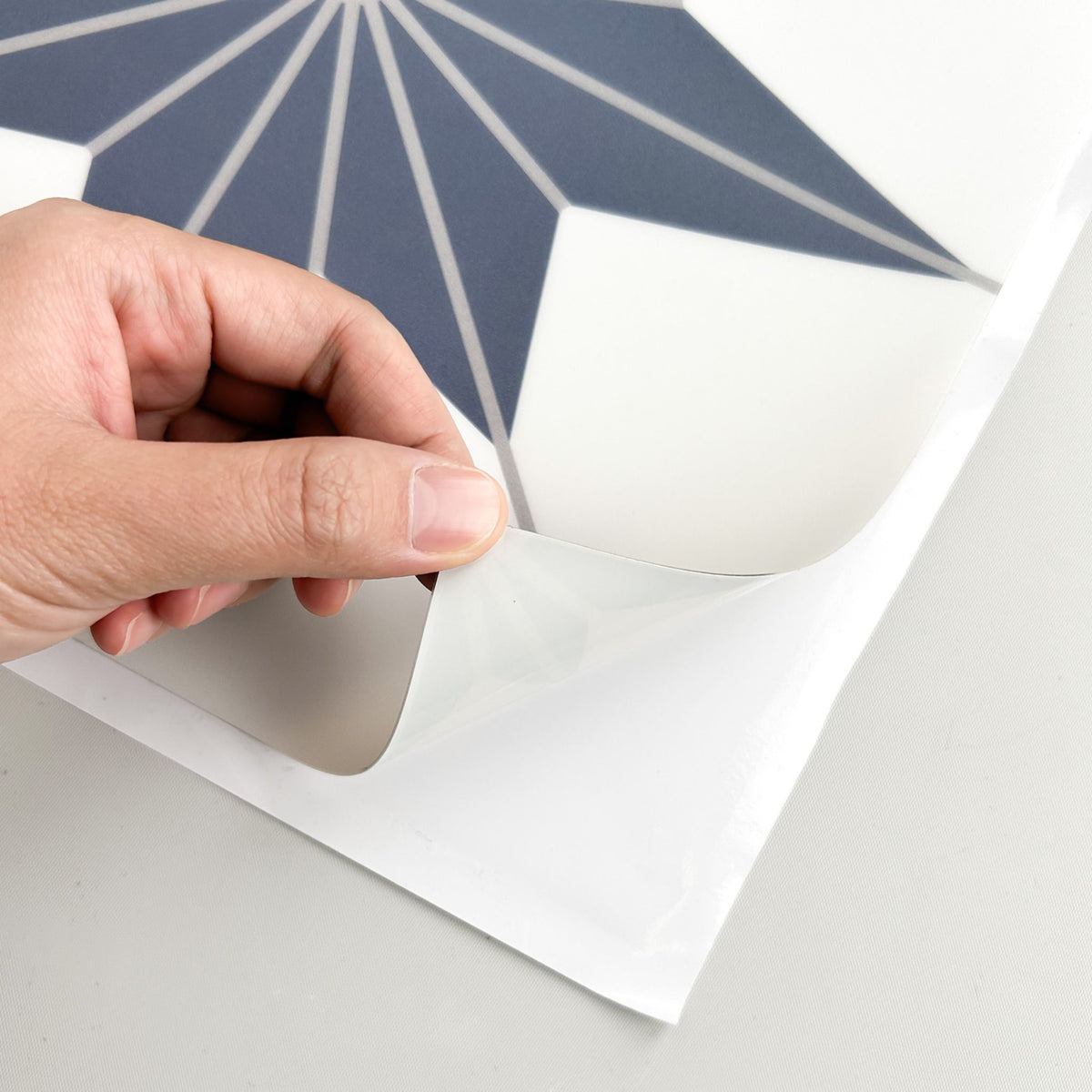 A close-up of a hand peeling back a clear protective film from a white sheet containing a navy blue and white geometric pattern.