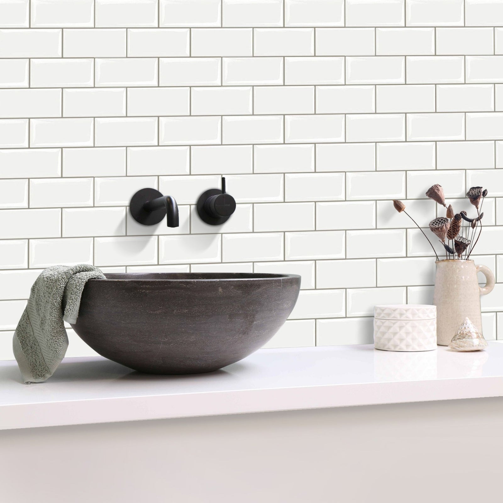 White subway tile backsplash on a bathroom wall with a dark charcoal vessel sink, matte black wall-mounted faucet, white countertop, and small decorative ceramic items.