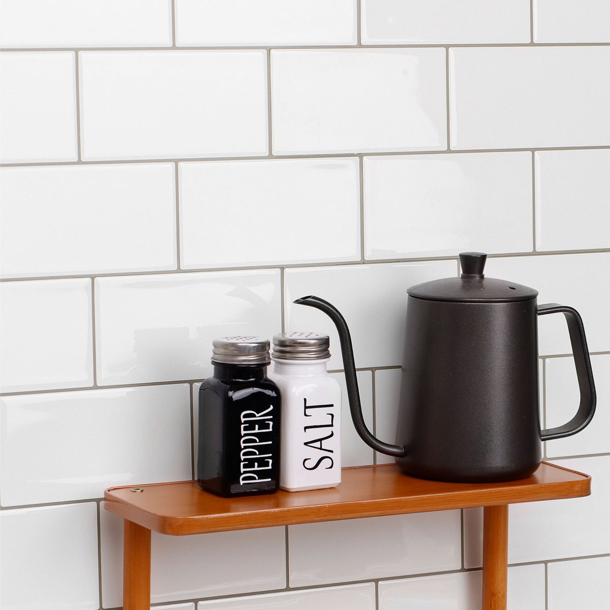 A matte black gooseneck kettle sits on a wooden shelf next to black and white pepper and salt shakers against a white subway tile wall.