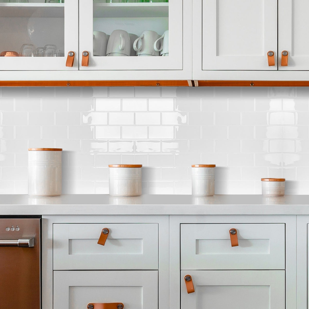 A styled kitchen scene with white cabinets, a white subway tile backsplash, and several white ceramic canisters with light wood lids arranged on the countertop.