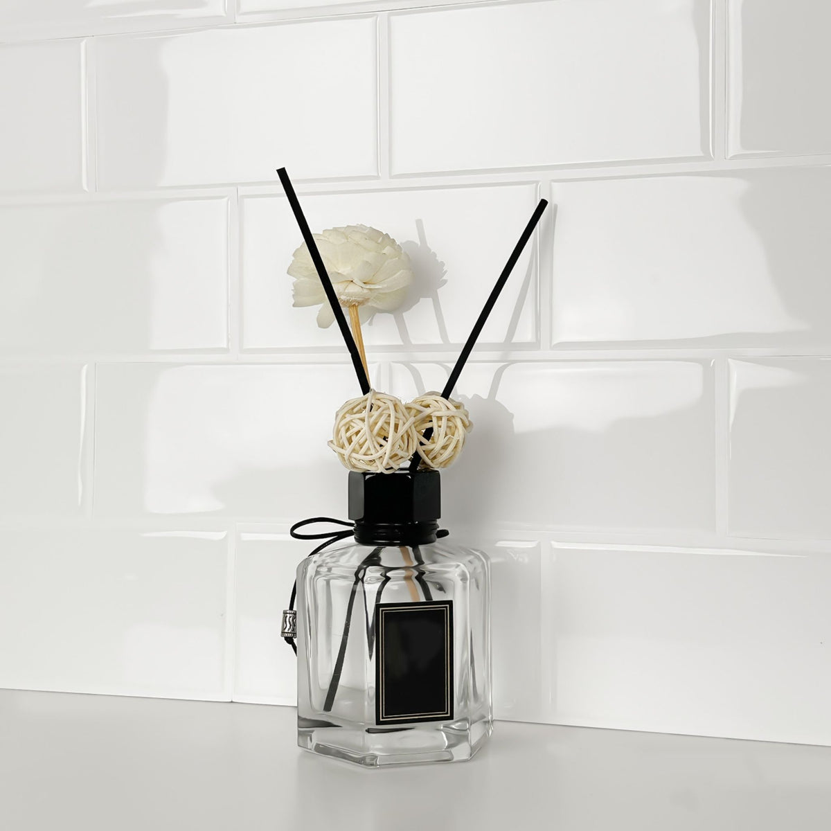 A clear glass reed diffuser bottle with a black cap and black reeds, decorated with a white flower and two woven beige spheres, set against a white subway tile background.