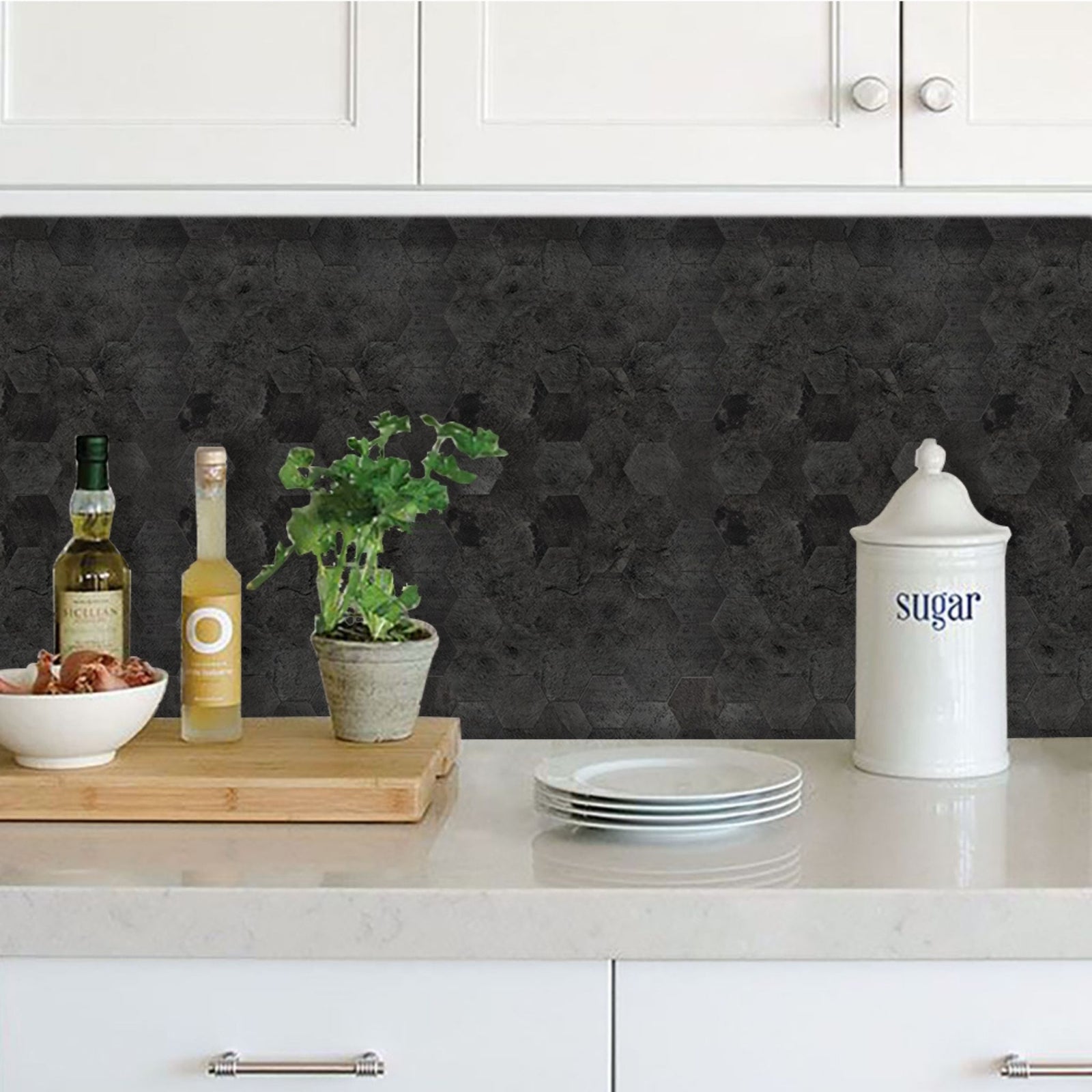 A kitchen countertop scene with a dark charcoal textured backsplash behind white cabinets. On the counter are stacked white plates, a white sugar canister, a small potted green plant on a wooden board, a bottle of olive oil, and a bottle of white wine.