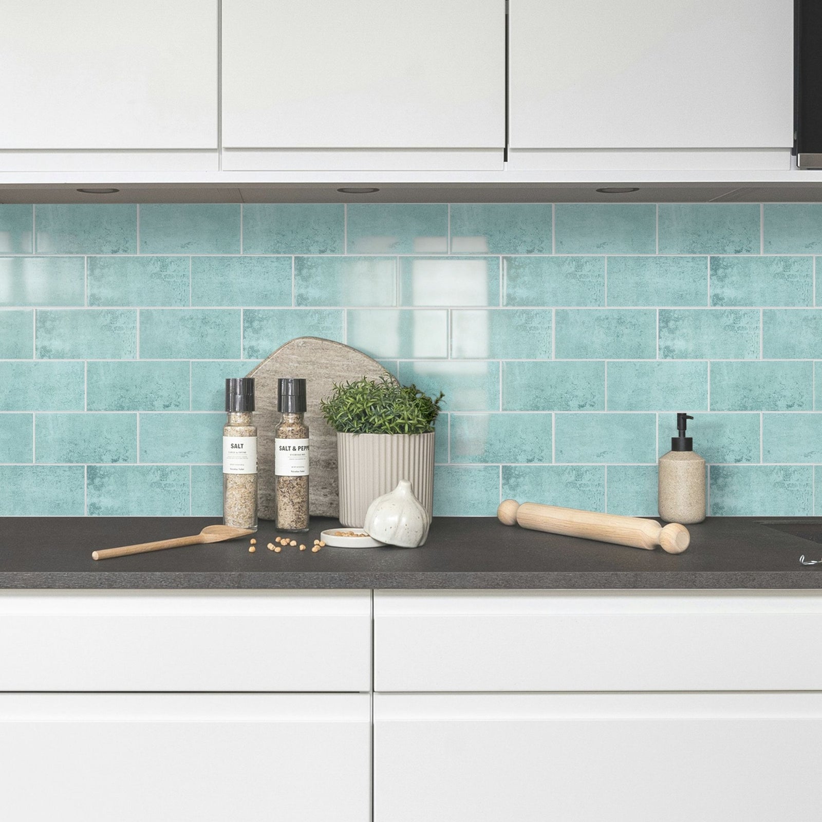 A modern kitchen scene featuring glossy light blue-green subway tiles on the backsplash, white cabinets, a dark countertop, and styled kitchen props including wooden utensils, spice jars, and a small potted plant.
