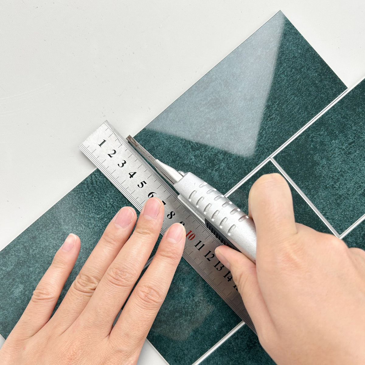 A close-up of a person's hands using a silver utility knife and metal ruler to measure and cut a glossy dark green tile sheet on a white surface.