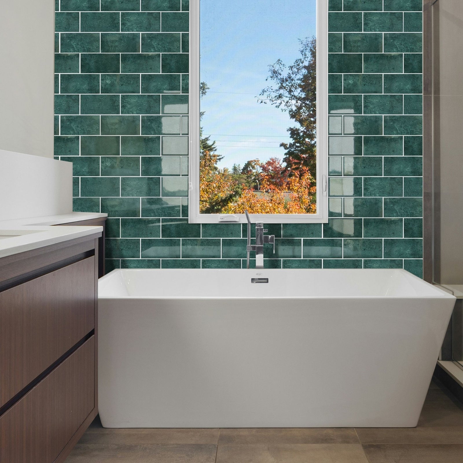 A modern freestanding white bathtub positioned in front of a green subway tile wall, with a window above showing an outdoor autumn view. A white countertop and dark wood vanity are visible on the left.
