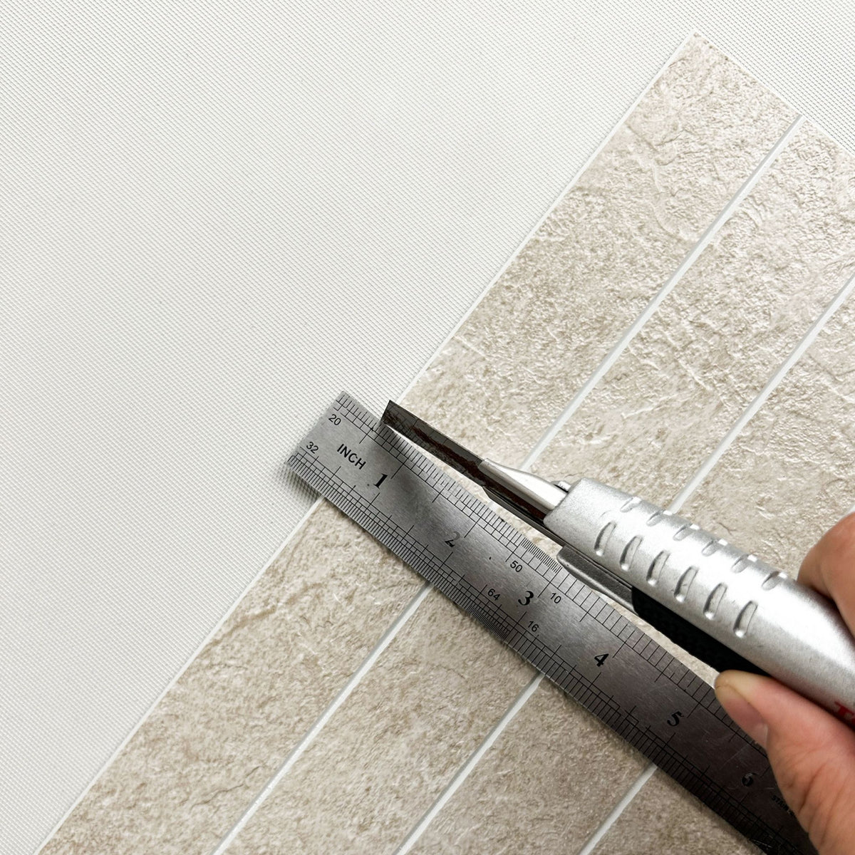 A close-up of a textured beige tile sheet being measured with a metal ruler and a utility knife against a white background.