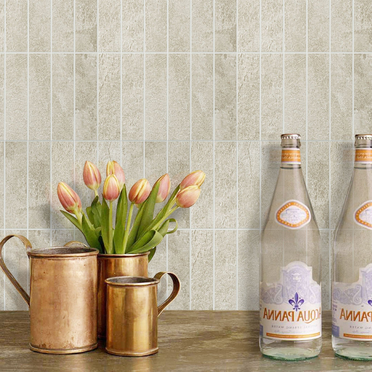 Stacked beige subway tiles on a wall behind three copper pots, one with tulips in and two water bottles. 