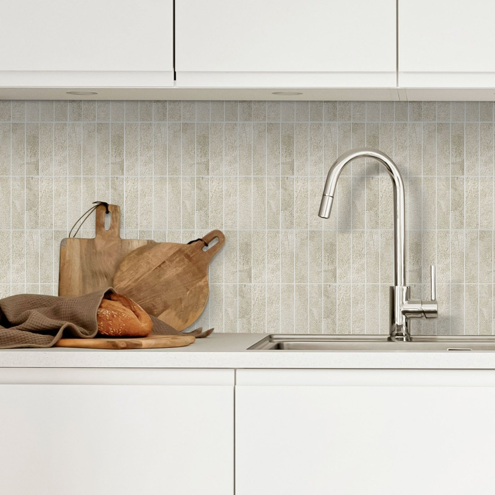 Stacked beige subway tiles as a backsplash in a white kitchen with a tap and wooden cutting boards and a loaf of bred on the counter.