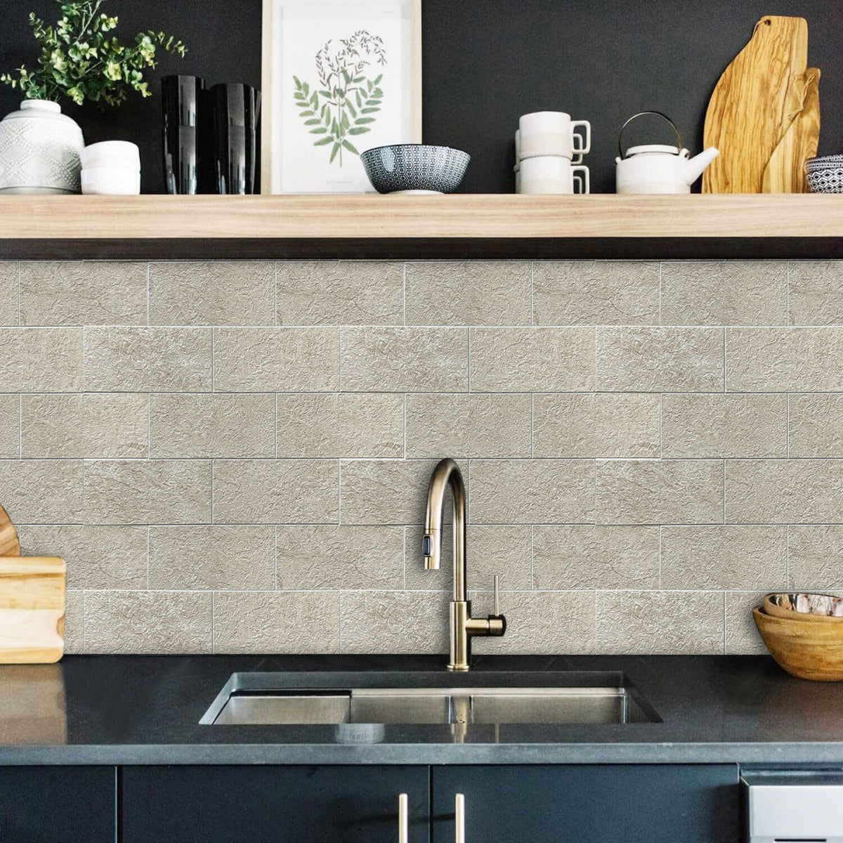 A kitchen backsplash made of light beige rectangular subway tiles installed behind a dark countertop and a brushed brass faucet. A wooden shelf above holds decorative items including mugs, a teapot, a framed print, and a vase.